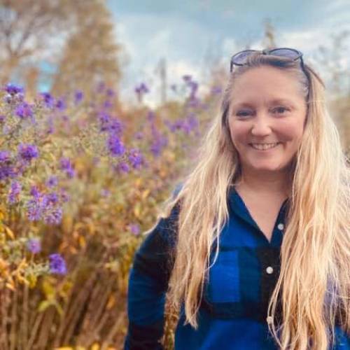 Photo of Jackie Herigodt with long blonde hair, smiling at the camera, standing in front of field of purple wildflowers.